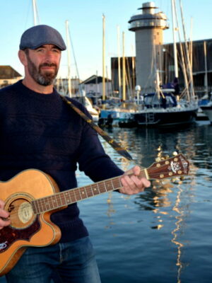 WIll Keating playing guitar in front of a harbour scene