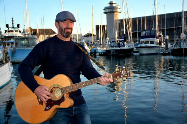 WIll Keating playing guitar in front of a harbour scene