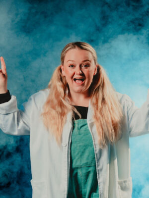 A blonde lady wearing a lab coat points two fingers in the air.