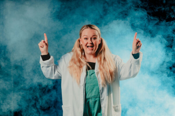 A blonde lady wearing a lab coat points two fingers in the air.