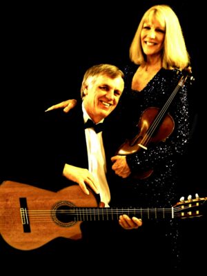 A man in a tuxedo and a lady in a sparkly dress stand close together holding their instruments - a classical guitar and a violin.