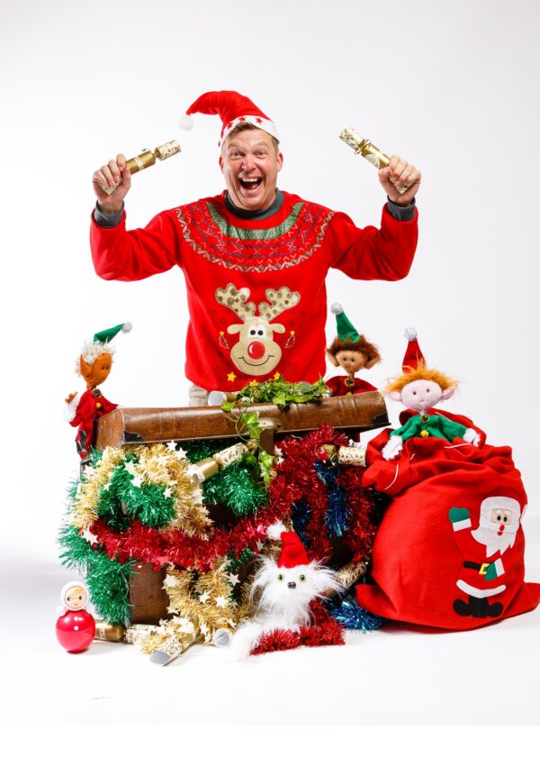 Craig dressed in a Christmas jumper and Santa hat, waves two crackers in the air, surrounded by Christmas presents and elves.