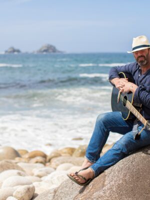 Will posing on a rock with his guitar. Sand and sea can be seen in the background.