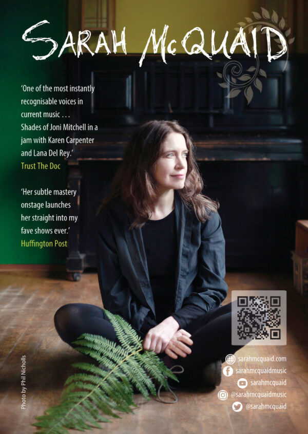 Sarah sits on a wooden floor in front of a piano whilst holding a fern leaf.