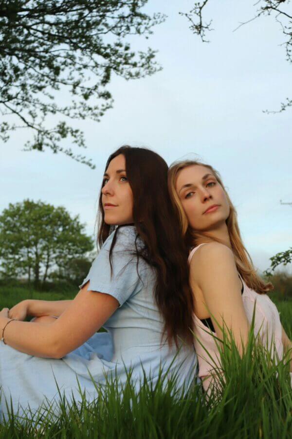 Amie and Chloe sit back to back in a field with trees in the background.