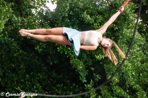 A circus performer hangs horizontally in the air, with the park greenery in the beackground.