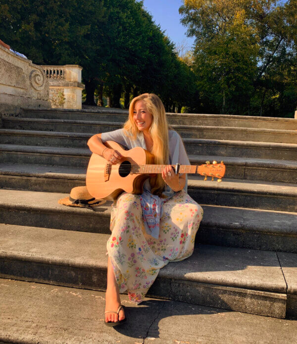 Suzy sits on stone steps, playing her guitar with a smile on her face.